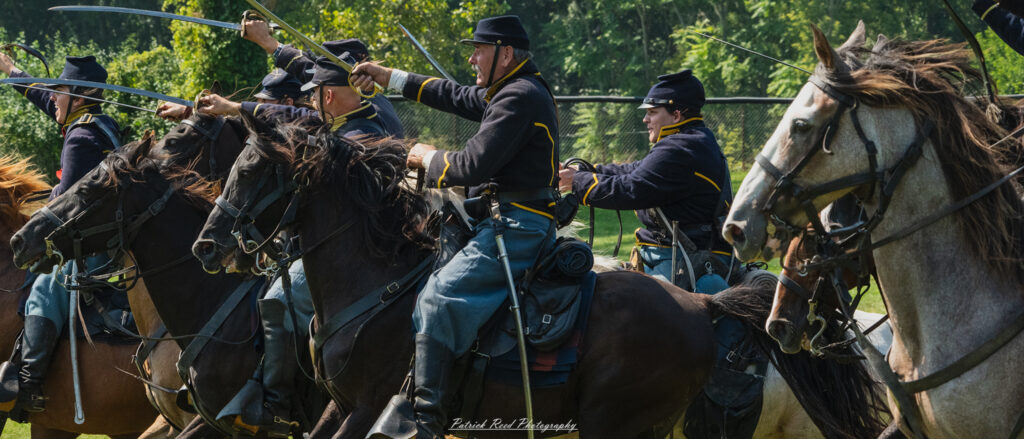 A group of cavalry soldiers charging forward on horseback, dressed in traditional military uniforms. Their expressions are fierce and determined as they gallop into battle, sabers drawn and weapons at the ready. The scene captures the energy and intensity of a cavalry charge, with dust and movement surrounding them.